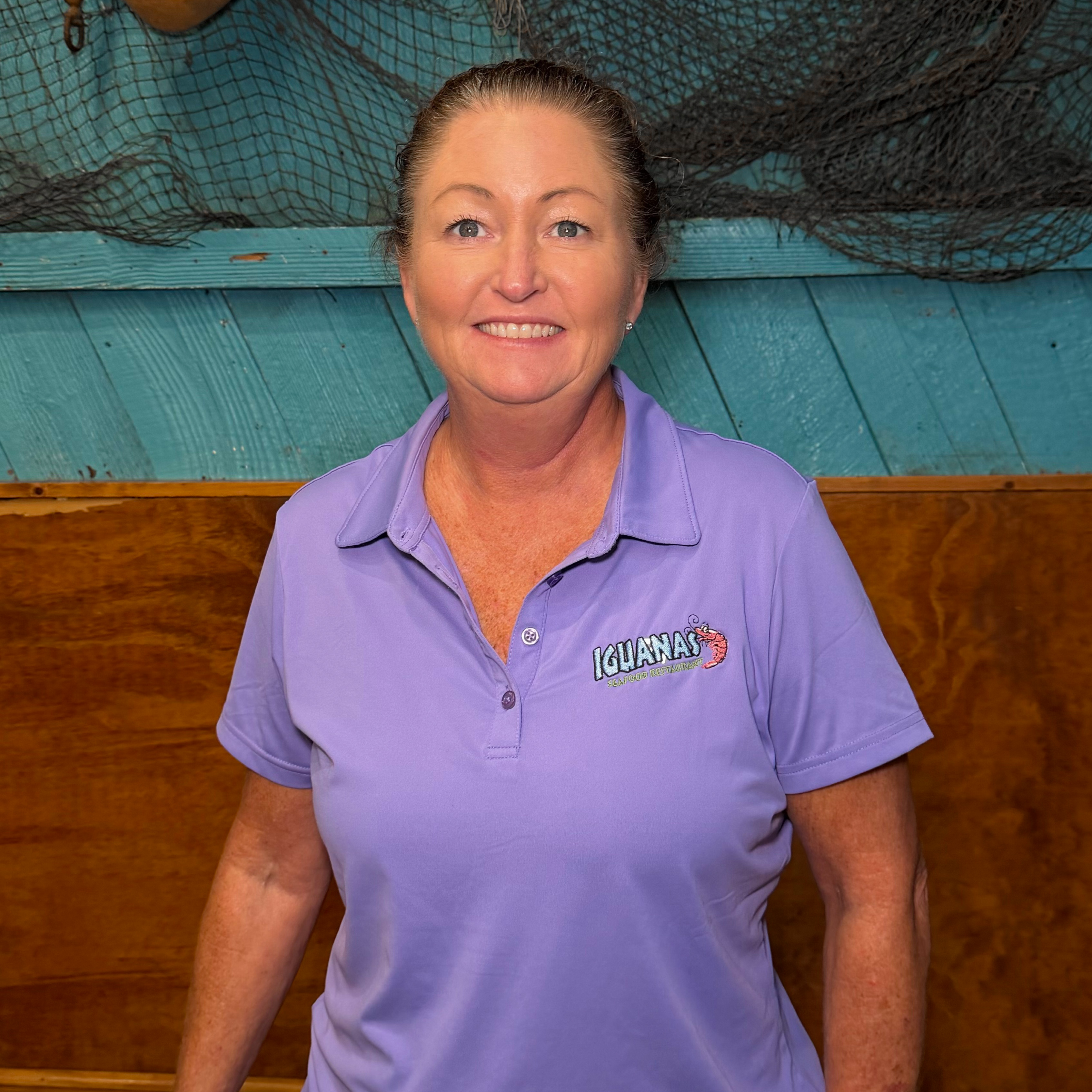Woman wearing a purple collared shirt with the Iguanas Logo stitched on the right side, with a turquoise and brown wall in the background with fishing paraphernalia. 