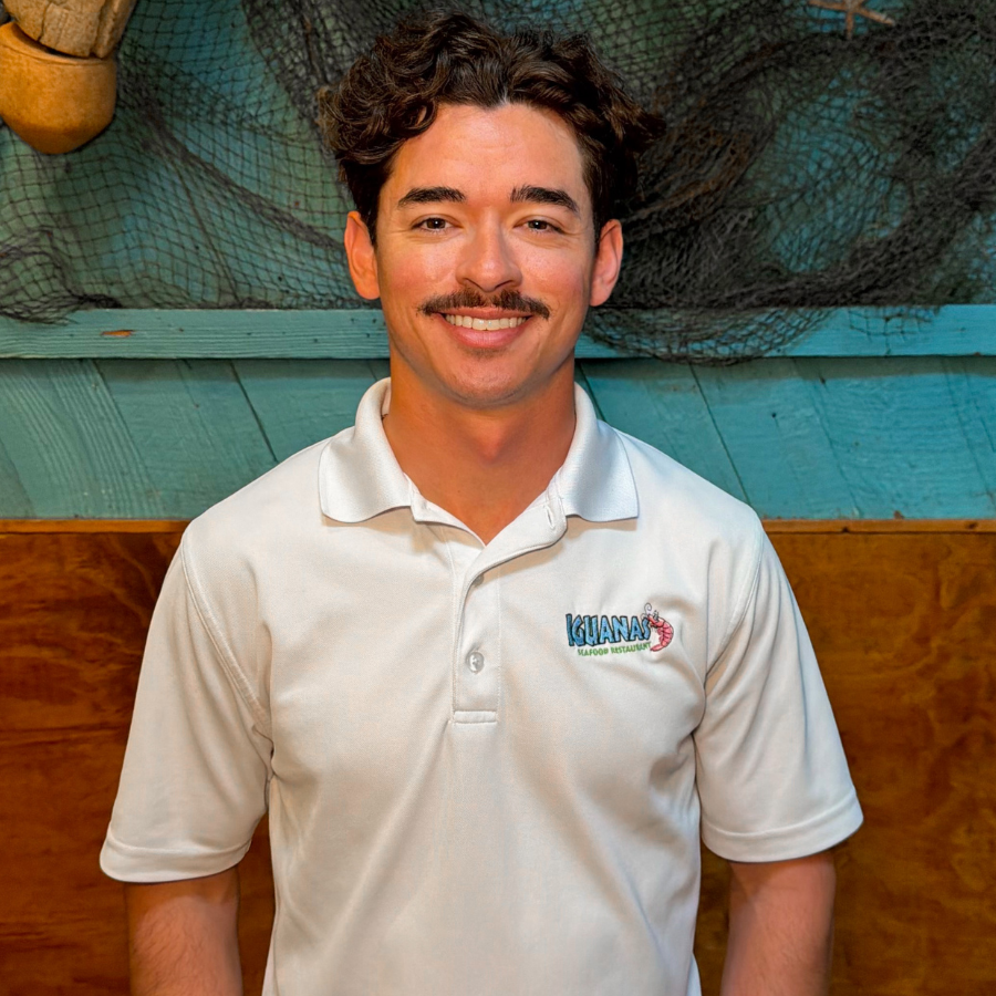 Man wearing a white collared shirt with the Iguanas Logo stitched on the front, with a turquoise wall and fishing paraphernalia in the background. 