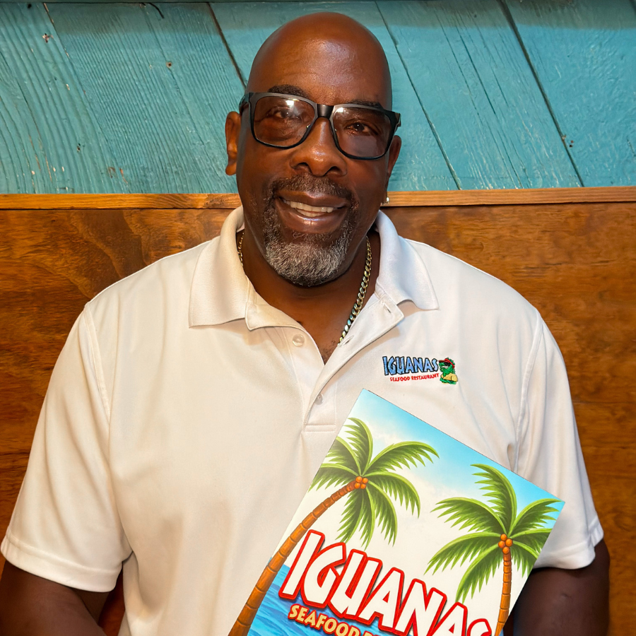 Man wearing a white collared shirt with the Iguanas Logo stitched on the front, with a turquoise wall and fishing paraphernalia in the background, holding an Iguanas Menu in his hands, displaying the front. 