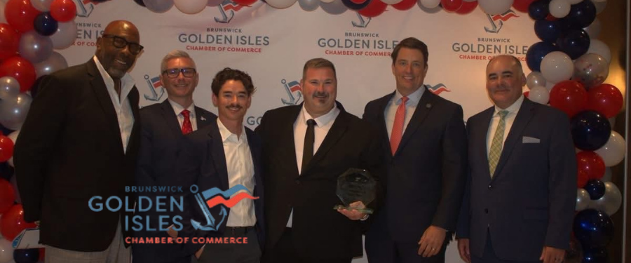 A photo of six men in front of a 'Golden Isles Chamber of Commerce' backdrop with balloons on either side, posing for a picture together holding the award, with the Brunswick Golden Isles Chamber of Commerce logo on the bottom left.