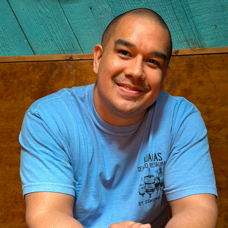 Man wearing a blue t-shirt with the Iguanas logo printed on the front, with a turquoise wall and fishing paraphernalia in the background. 